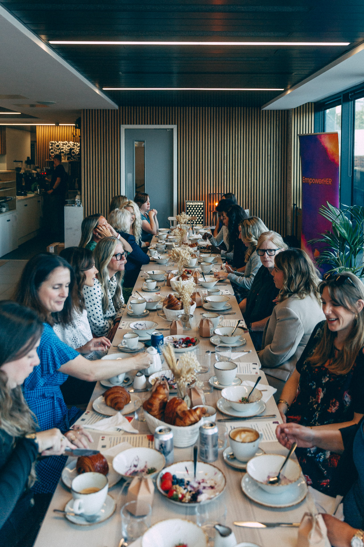 A long breakfast table of women at an EmpowerHER gathering, with the EmpowerHER banner